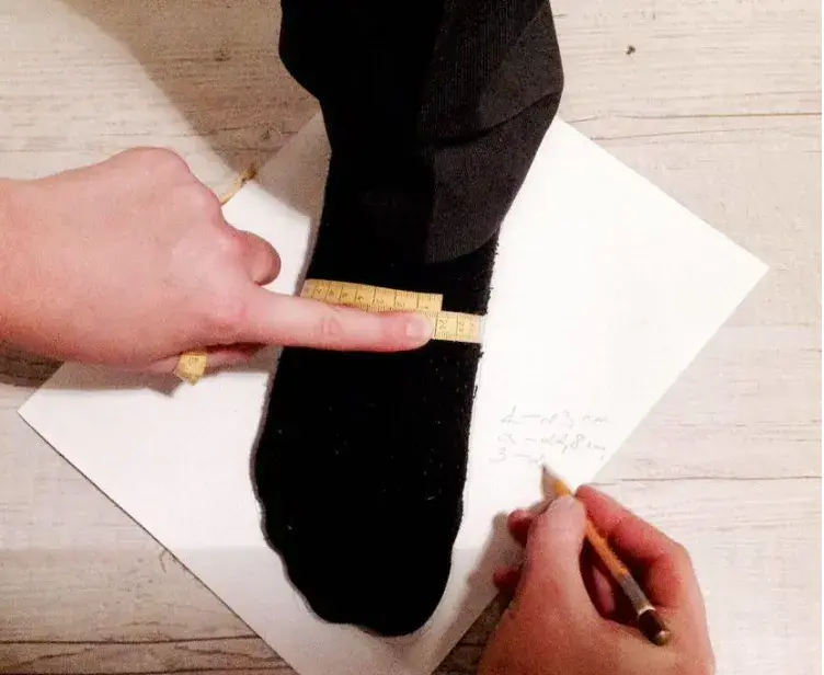 A tape measure around the instep of the foot, and a hand noting measurements on the paper