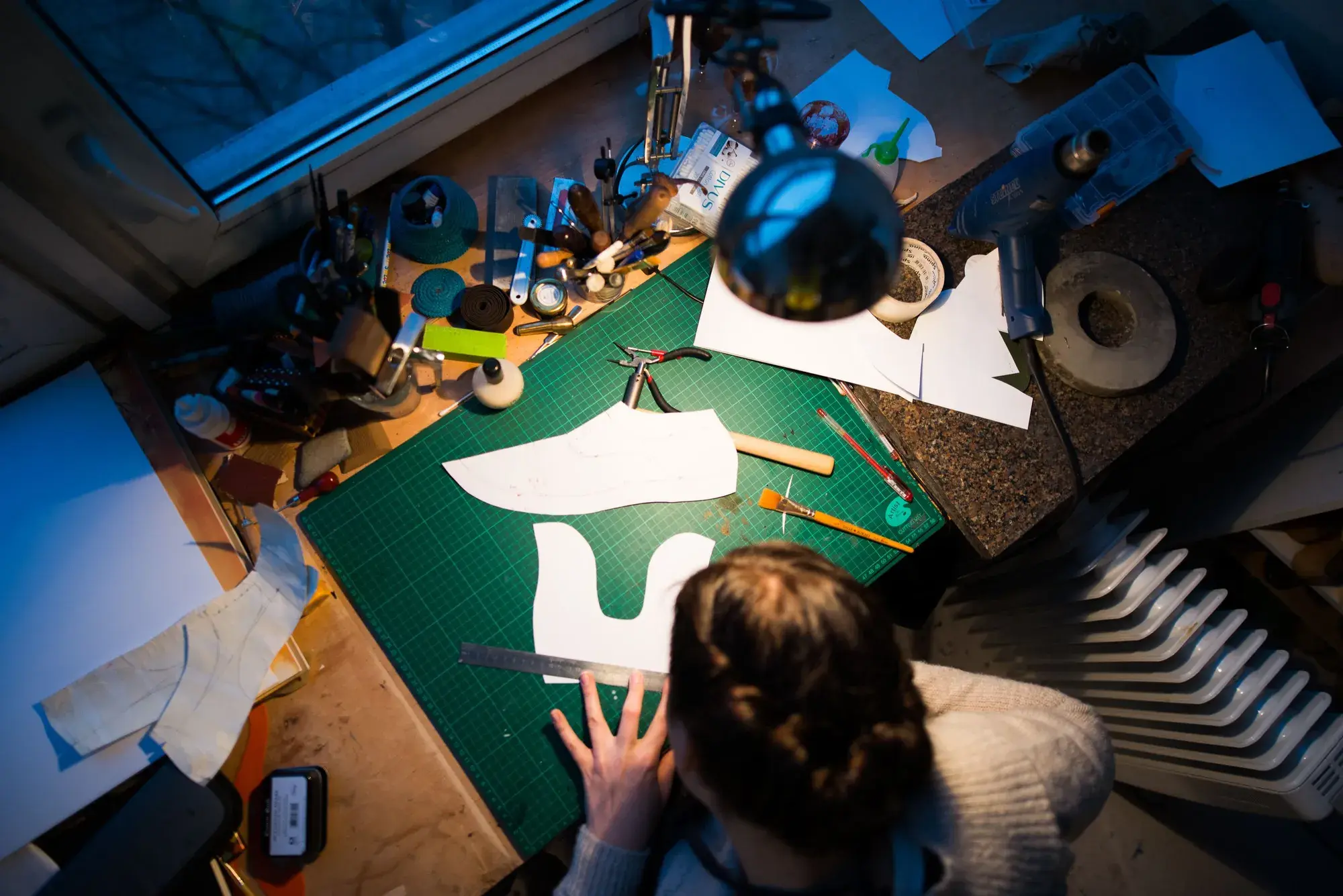 Overhead view of the bench: cut white pattern pieces of a shoe upper laid on the green cutting mat under a lamp