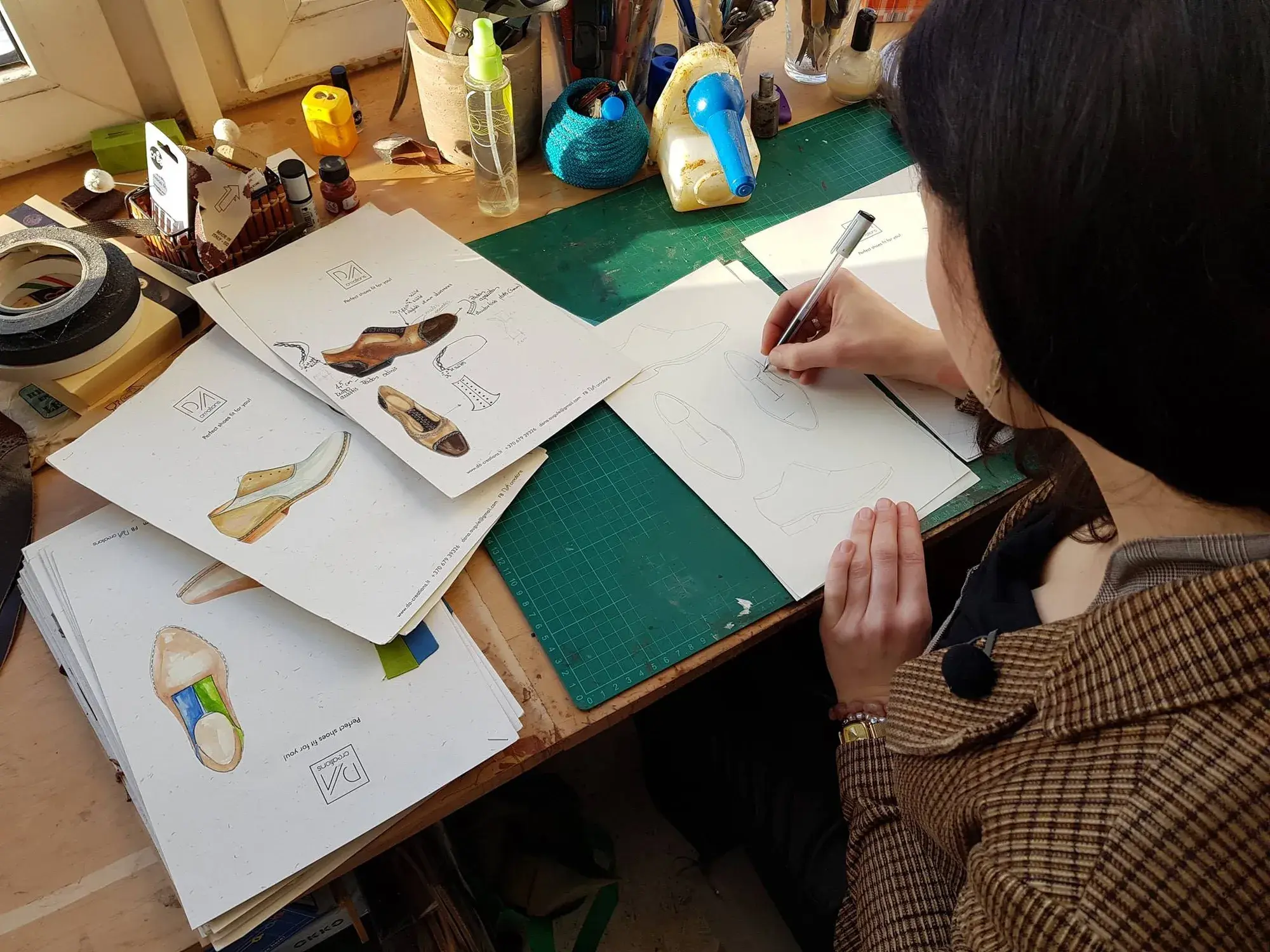 The maker drawing shoe designs in pen, watercolour sketches of finished models spread across the bench
